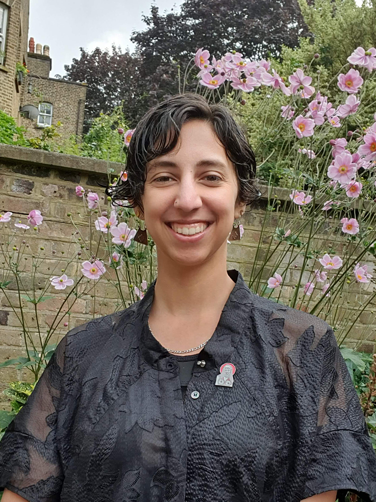 A photograph of Maka in a garden. They smile at the camera while standing in front of tall pink flowers. They have short dark curly hair and wear a black patterned see through shirt with a black and red drag queen pin.