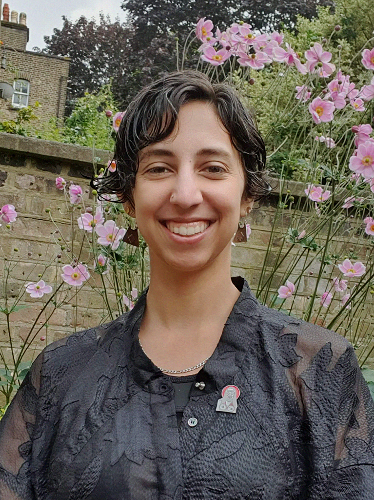 A photograph of Maka in a garden. They smile at the camera while standing in front of tall pink flowers. They have short dark curly hair and wear a black patterned see through shirt with a black and red drag queen pin.